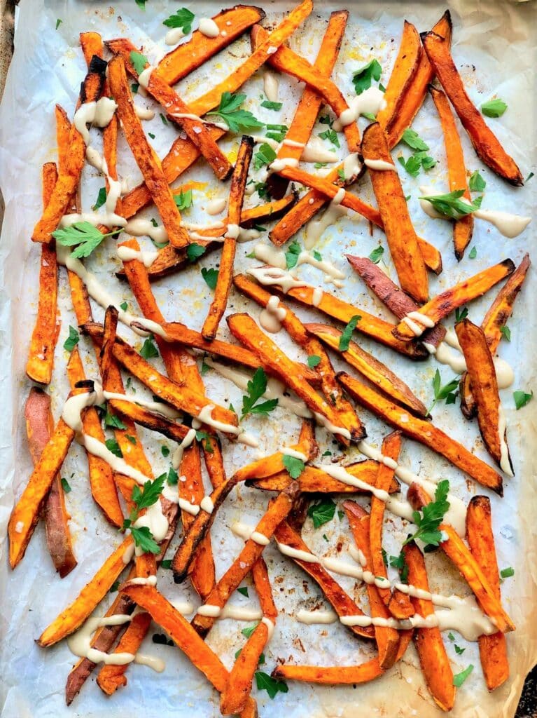 Overhead shot of the cooked sweet potato fries on a lined baking sheet drizzled with lime and tahini dressing and garnished with fresh cilantro. 