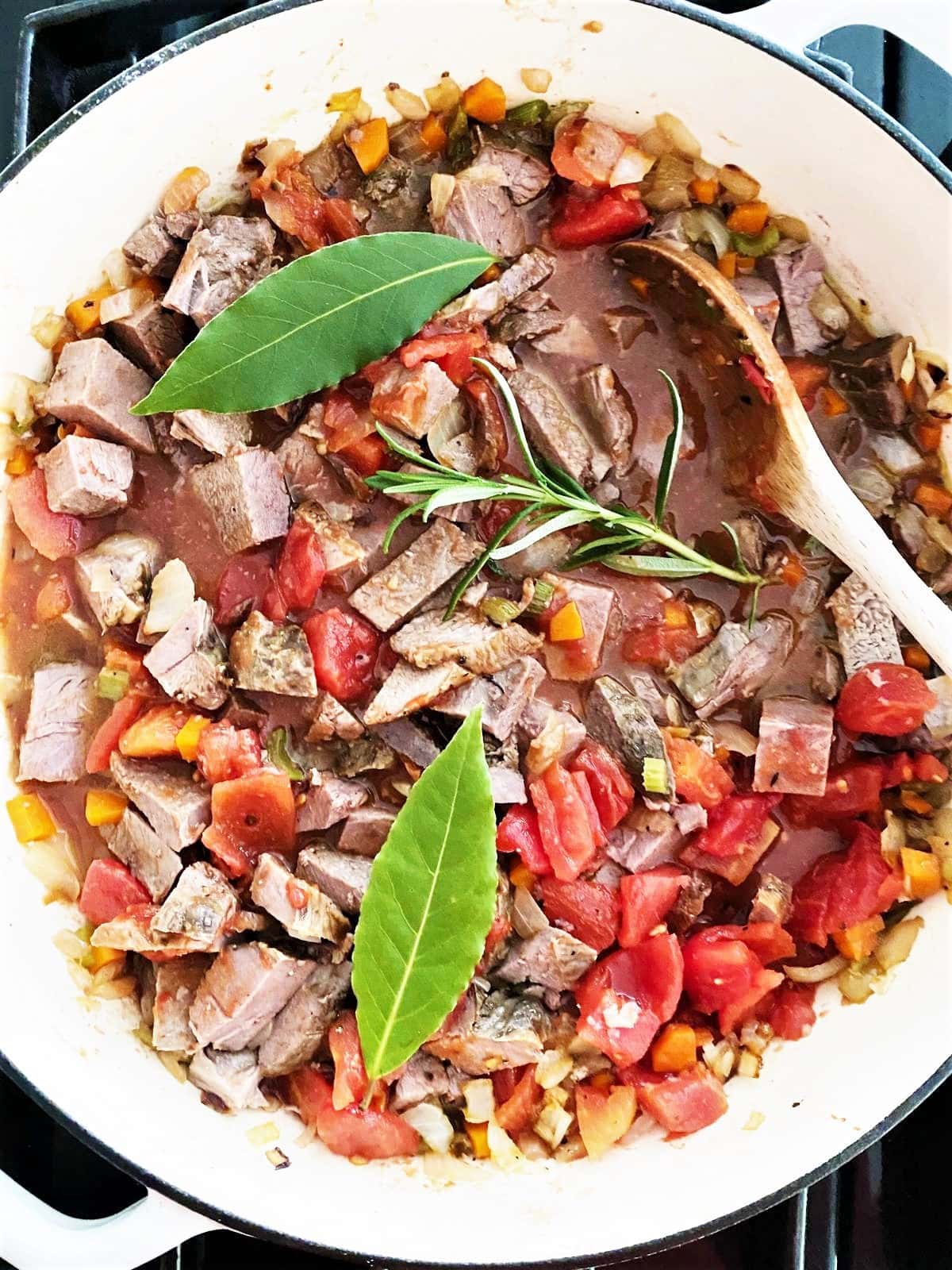 Top down image of large pot with part cooked lamb ragu topped with a sprig of fresh rosemary and two fresh bay leaves.