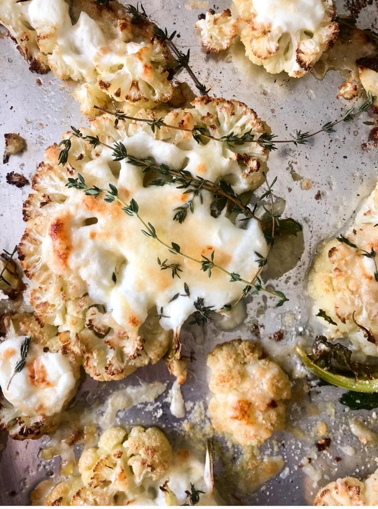 Top down image of roasted cauliflower steaks topped with creme fraiche, shredded cheese and sprigs of thyme on a roasting tray.