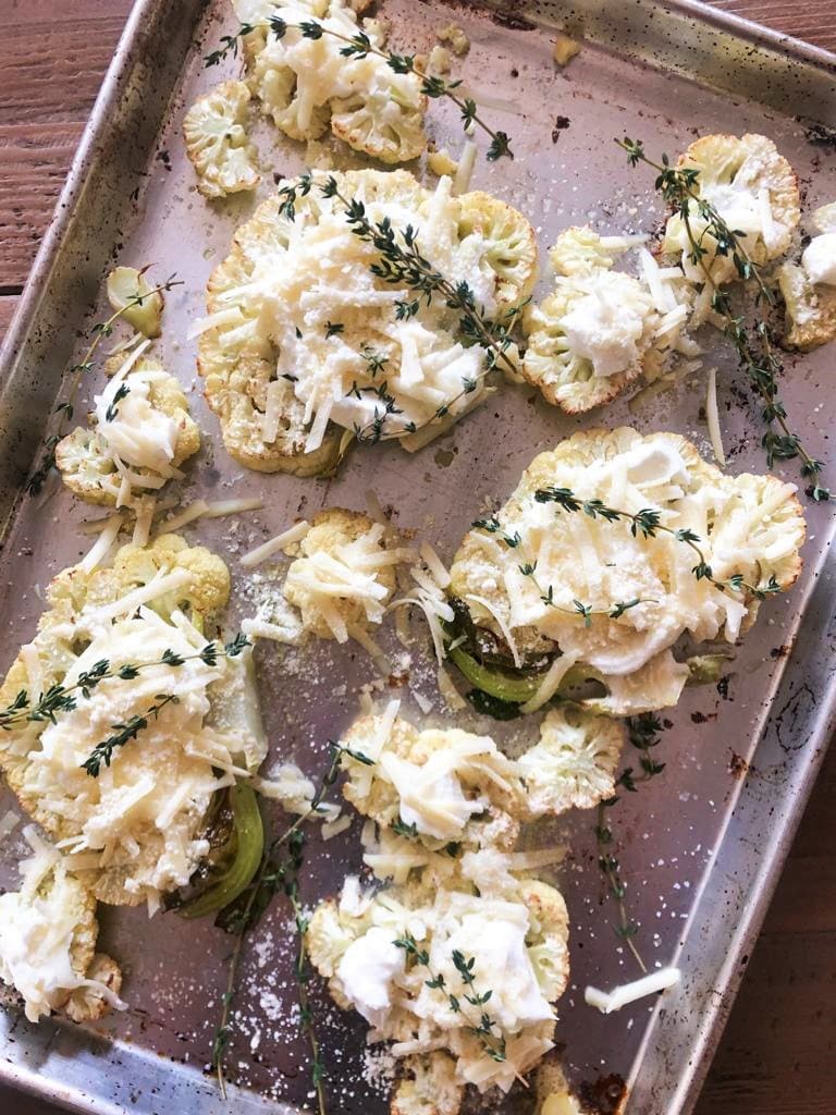 Flatlay image of roasted cauliflower steaks on a roasting tray topped with creme fraich, shredded cheese and sprigs of time, ready to go into oven.
