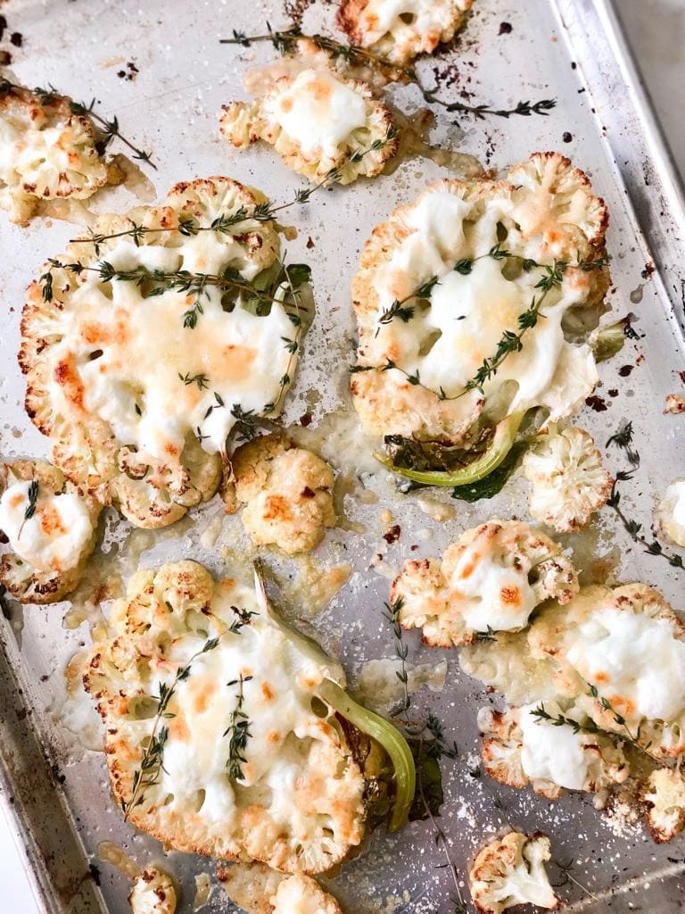 Top down image of roasted cauliflower steaks topped with creme fraiche, shredded cheese and sprigs of thyme on a roasting tray.