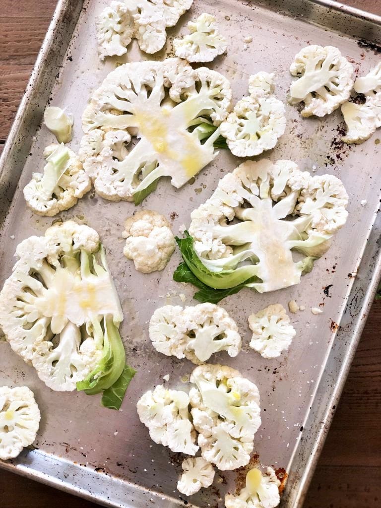 Flatlay image of roasting tray with cauliflower steaks and florets, drizzled with olive oil.