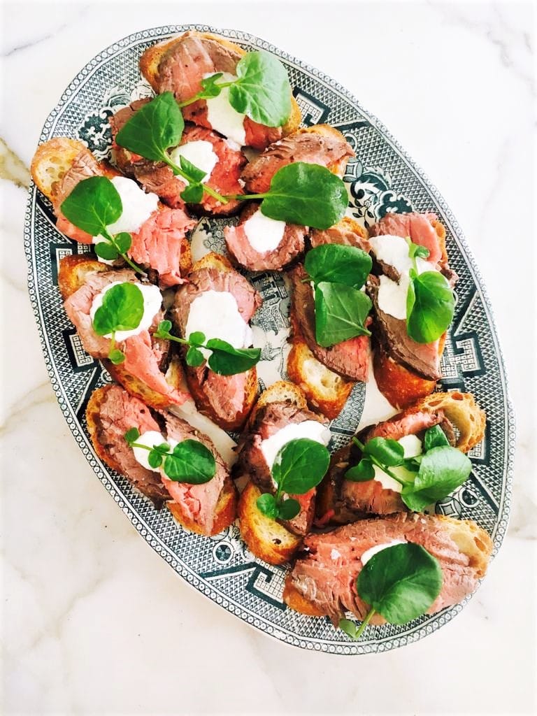 Oval platter of steak crostini shown from above including horseradish and sour cream sauce and watercress garnish.  