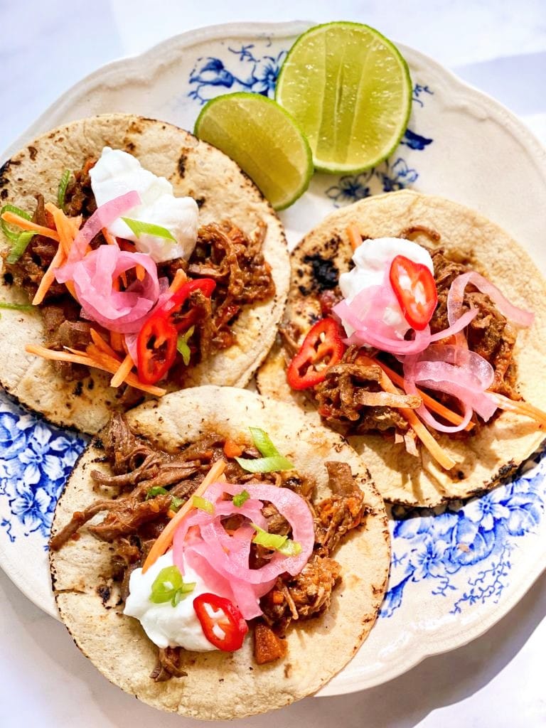 Top down image of blue and white plate with 3 flour tortillas topped with pulled pork, pink pickled onion, sliced red chili and cilantro and some wedges of lime.