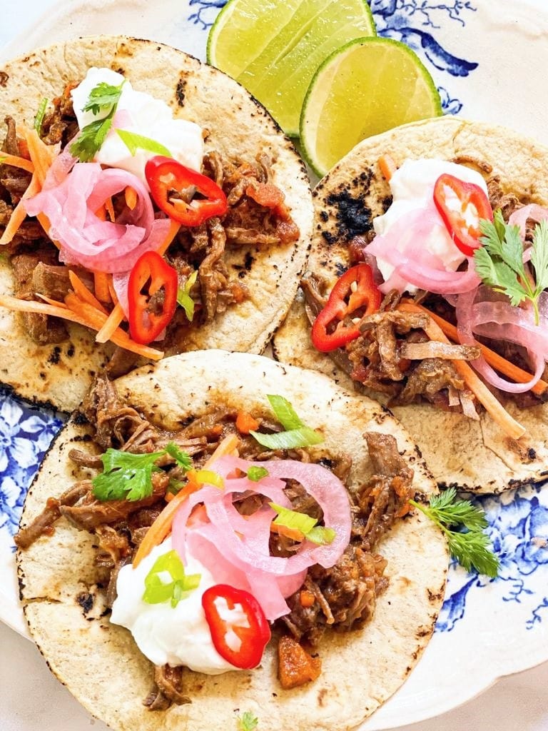 Top down image of blue and white plate with 3 flour tortillas topped with pulled pork, pink pickled onion, sliced red chili and cilantro and some wedges of lime.