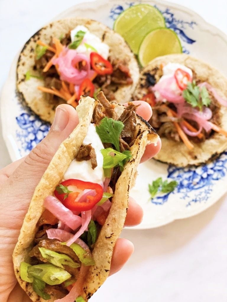 Image of hand holding a taco filled with pulled pork, pink pickled onion, sliced red chili and cilantro and a blue and white plate with two further tacos in the background.