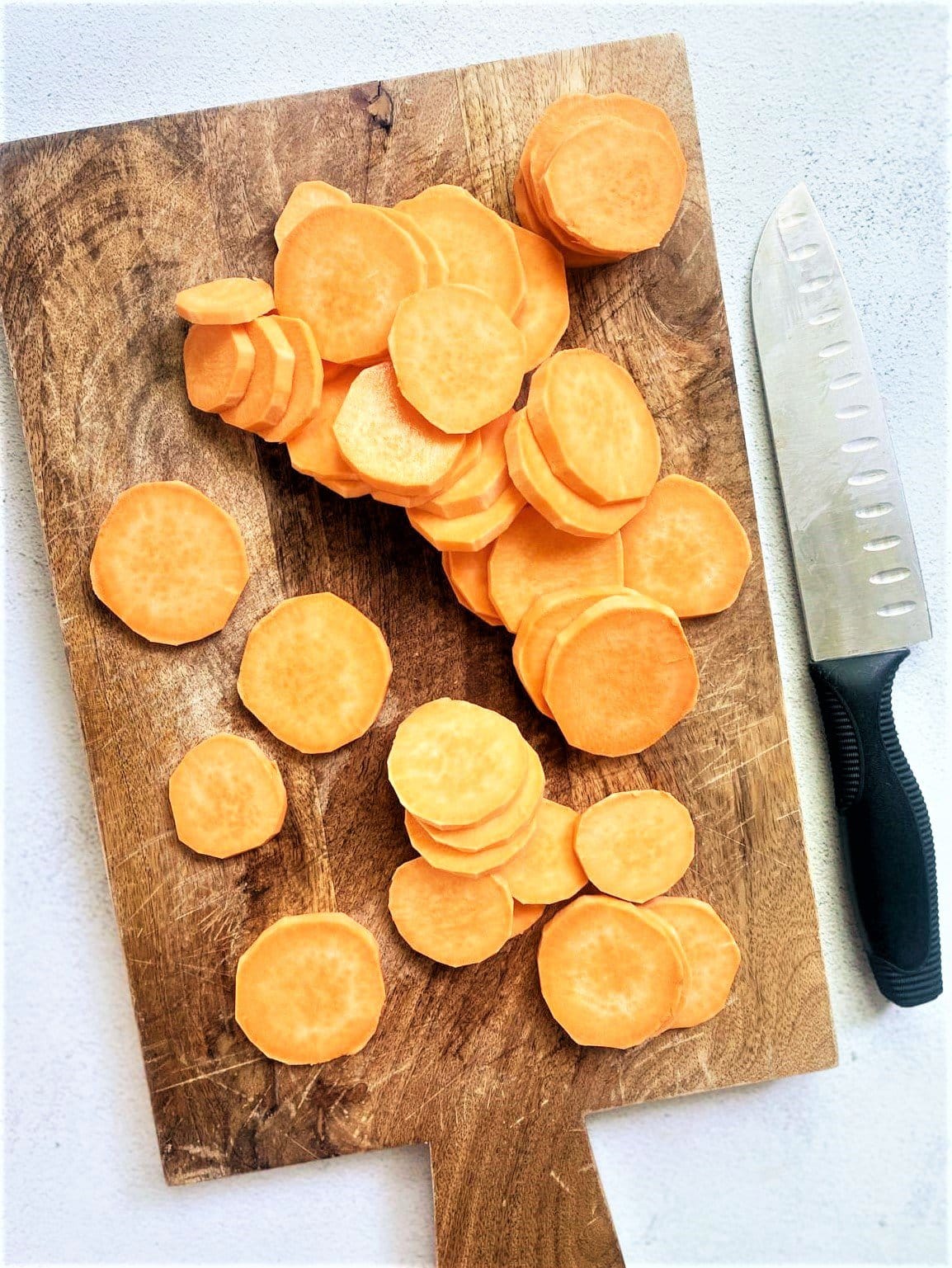 Top down image of sweet potatoes slices on top of a wooden board with a kitchen knife alongside.