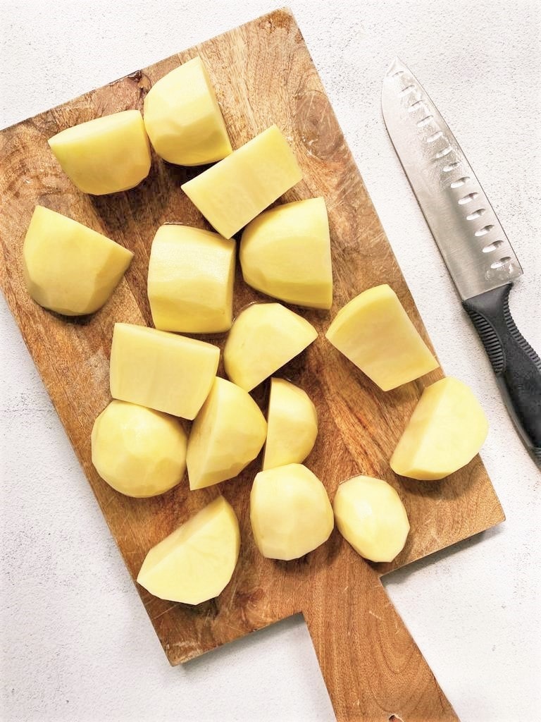 Top down image of wooden board with peeled and chopped potatoes and a kitchen knife set alongside.