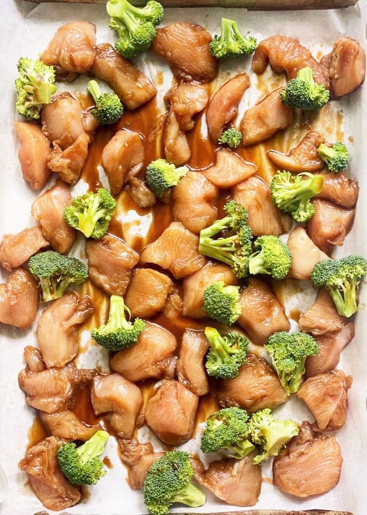 Top down image of sheet pan with marinated chicken and raw broccoli florets.