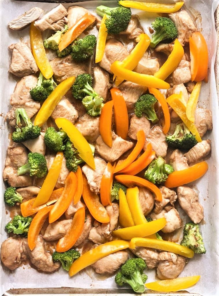 Top down image of sheet pan of part cooked chicken and broccoli florets and raw slices of yellow and orange bell peppers scattered over.