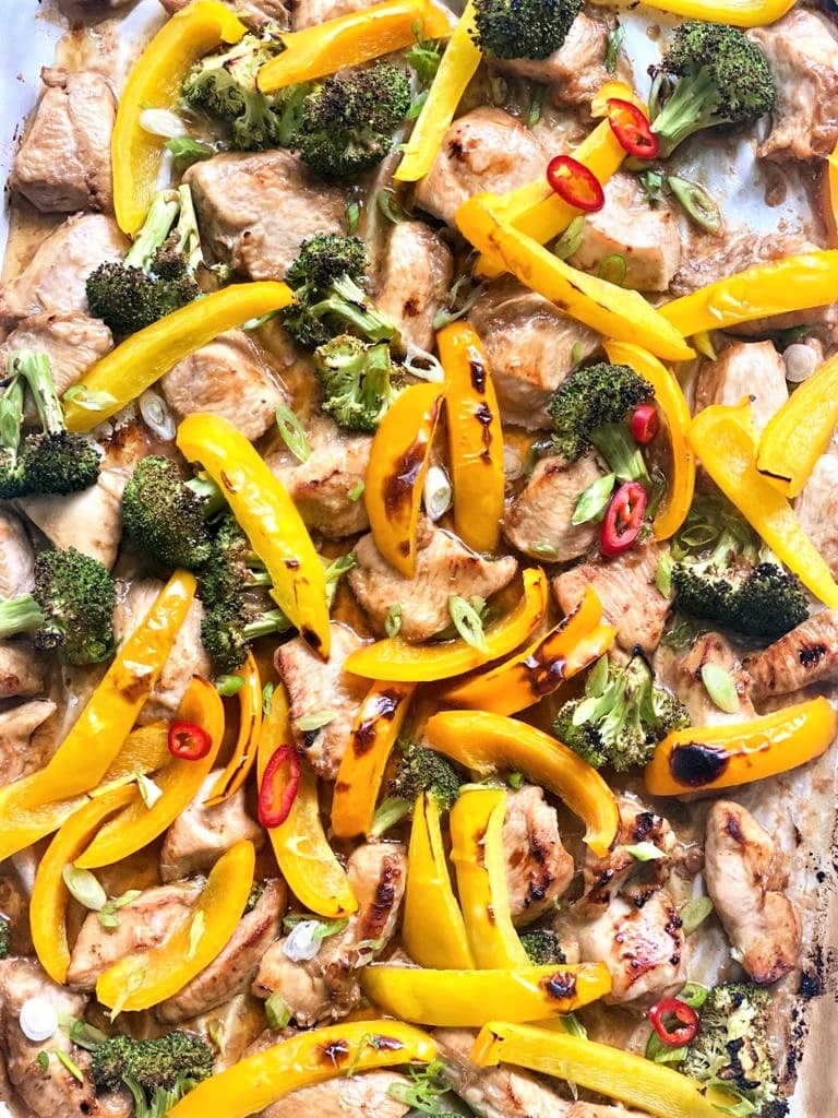 Top down image of sheet pan of part cooked chicken and broccoli florets and sliced yellow and orange bell peppers garnished with sliced red chili and green onion.