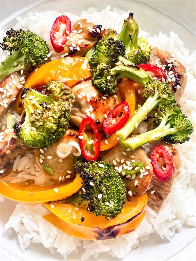 Top down image of a white bowl filled with steamed white rice and topped with chicken, broccoli and bell pepper stir fry and garnished with green onions, red chili and sesame seeds.