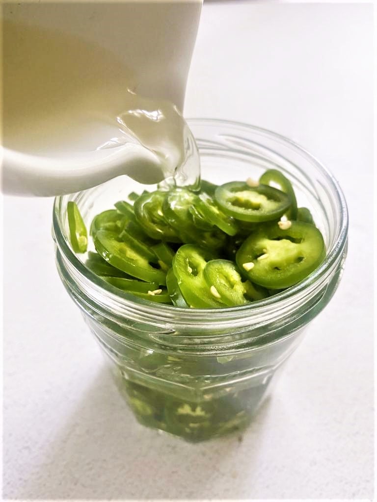 A jar of sliced green Serrano peppers with a jug of pickling liquor being poured over.