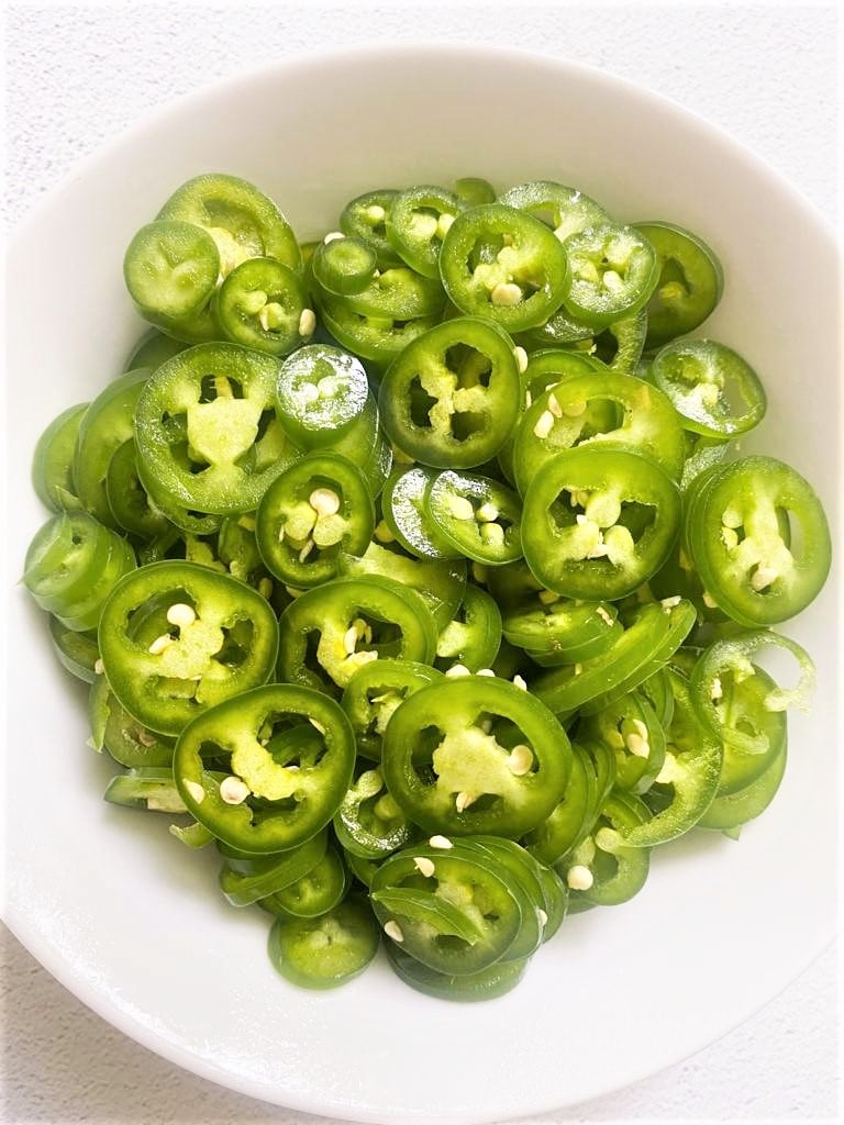 A top down image of a while bowl full of sliced green Serrano peppers.