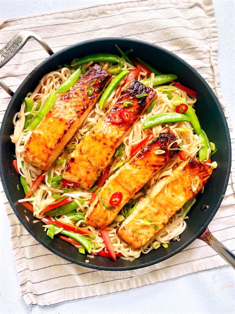 Top down image of skillet set on a kitchen towel. Skillet is filled with noodles, stir fried vegetables and four teriyaki salmon fillets, garnished with sliced red chilies and sesame seeds.