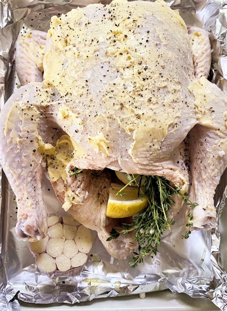 Top down image of whole raw chicken set into a roasting dish,with softened butter spread across the bird, lemon and thyme in the cavity and a bulb of garlic set into the dish.