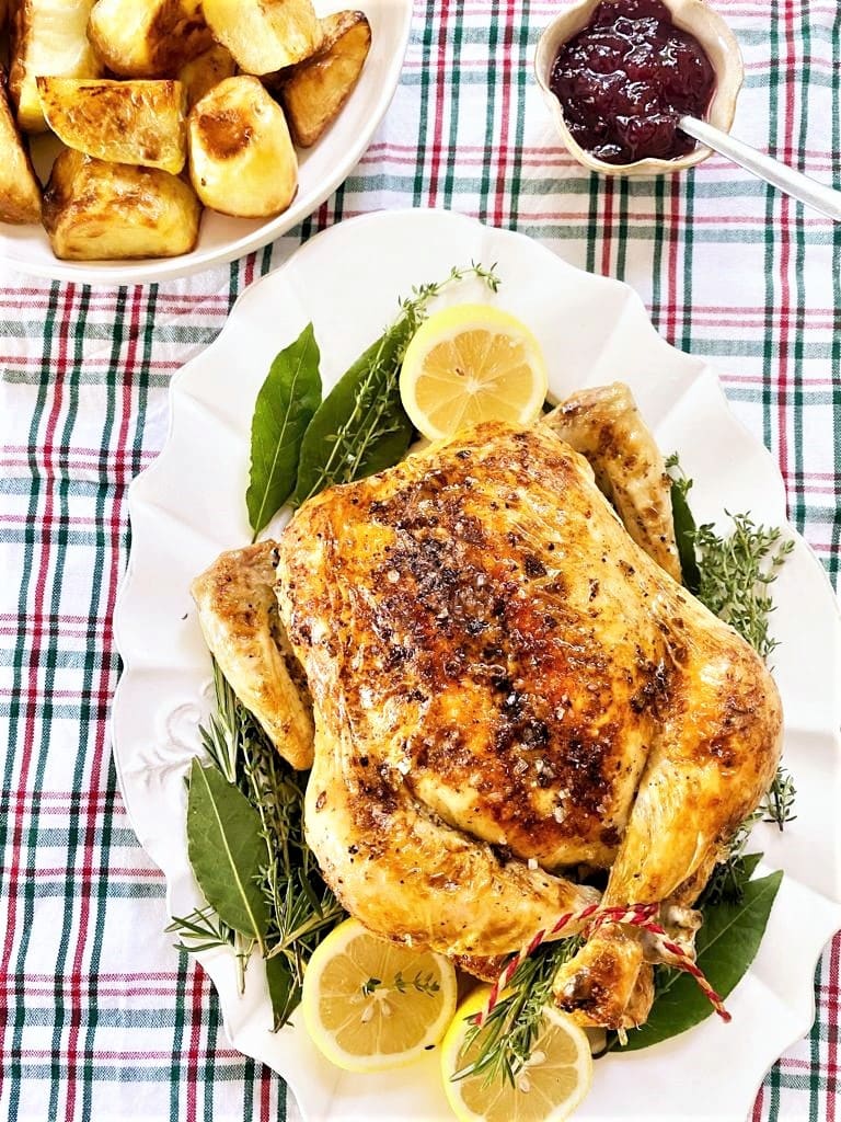 Top down image of whole roast chicken, with lemon, fresh thyme and bay leaves and sides of crispy roast potatoes and cranberry sauce.