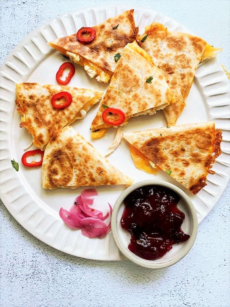 Top down image of leftover turkey and cheese quesadillas garnished with red chili, pickled red onions and a bowl of cranberry sauce