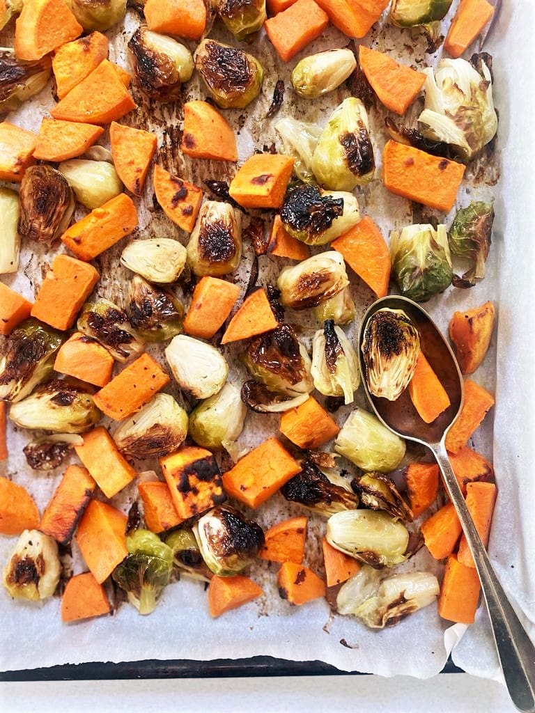 Close up image of baking sheet lined with baking paper containing roasted brussel sprouts and sweet potato with a serving spoon