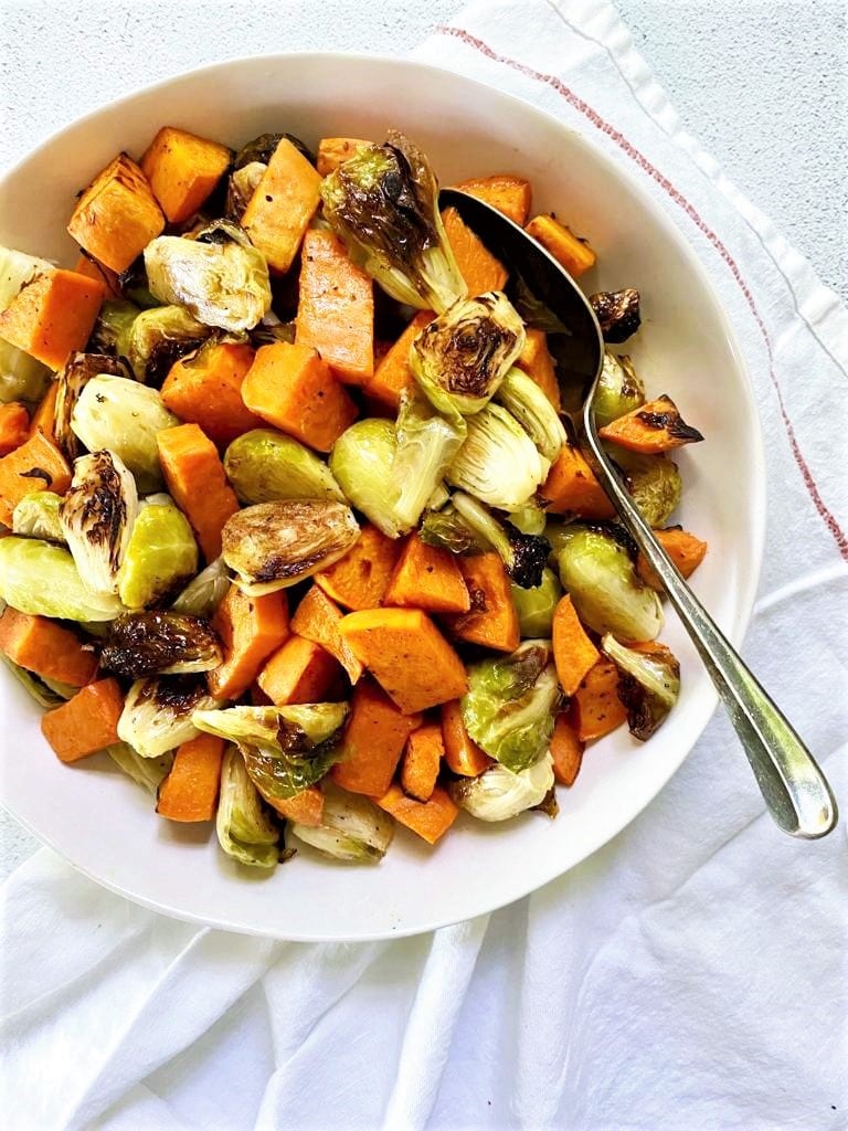 White bowl containing roasted brussel sprouts and sweet potato with a serving spoon