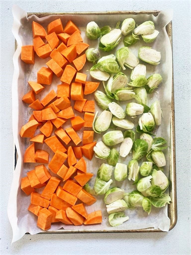 Top down image of baking sheet lined with baking paper one half of tray containing raw sweet potato, the other half raw brussel sprouts