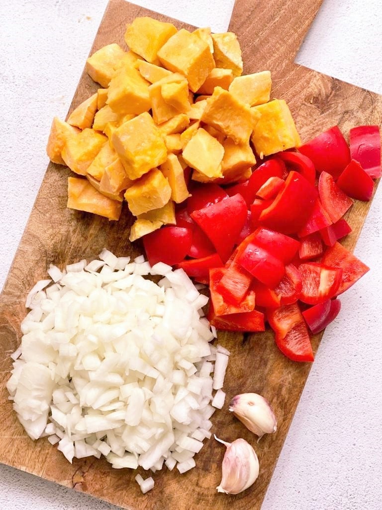 Chopped butternut squash, red bell pepper, onions and 2 whole cloves of garlic, set on a wooden chopping board 