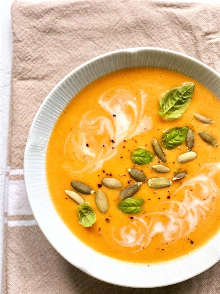 Top down image of white bowl filled with butternut squash and red pepper soup, topped with a swirl of coconut milk, sunflower seeds and fresh herbs