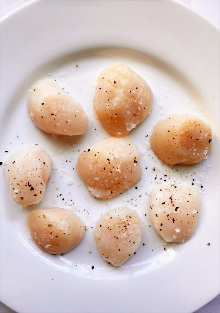 White plate with raw scallops seasoned with sea salt and black pepper.