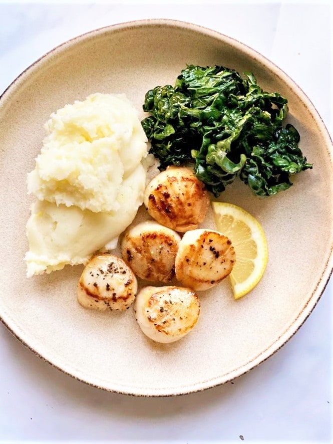 Top down image of plate of pan seared scallops in butter with a slice of fresh lemon and sides of mashed potato and steamed kale.