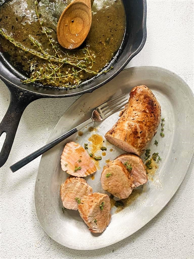 Serving platter with sliced roasted pork tenderloin with a butter and thyme pan sauce and cast iron skillet with butter pan sauce