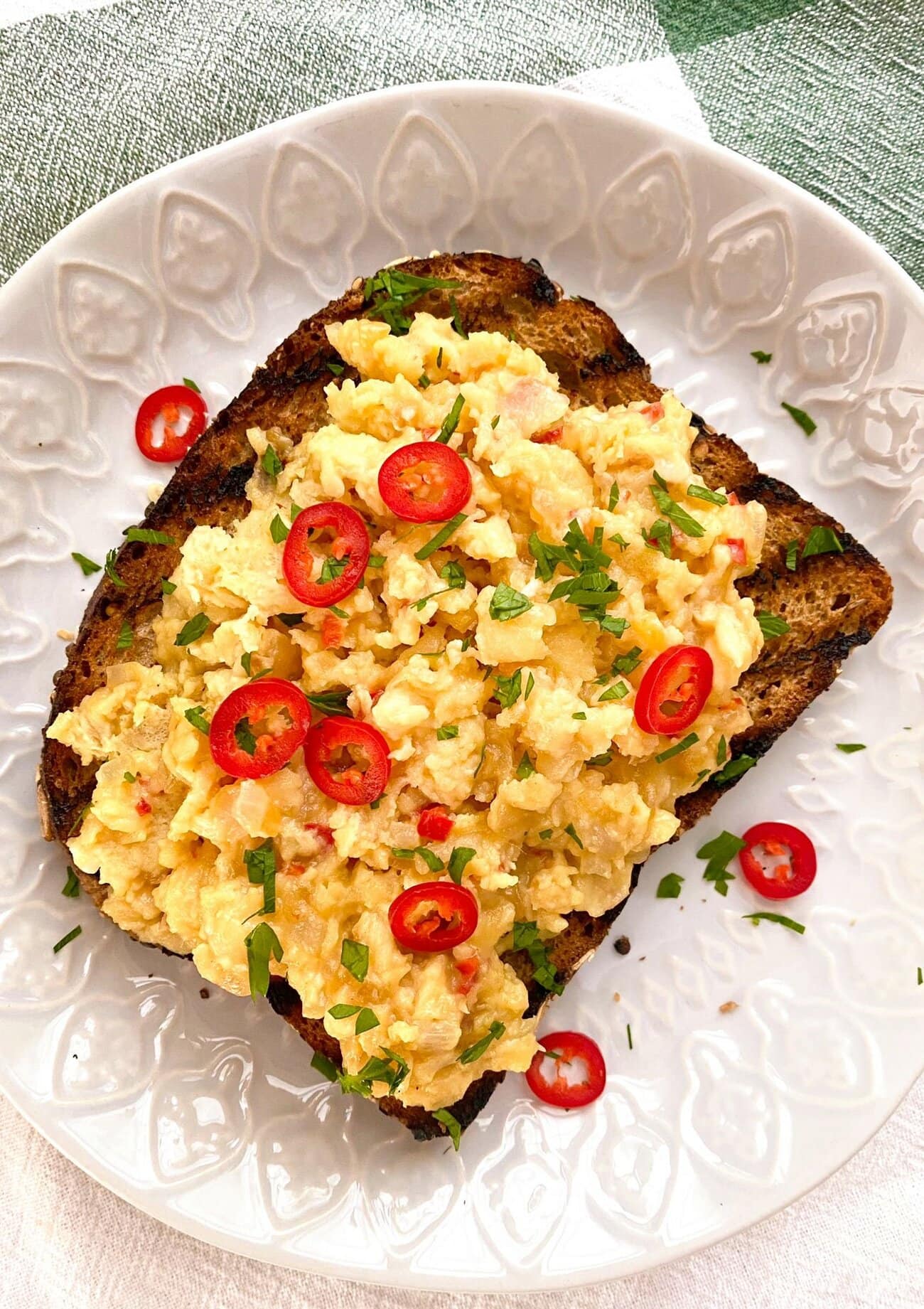 White plate with a slice of toasted granary bread topped with chili scrambled eggs and garnished with slices of red chili and freshly chopped parsley