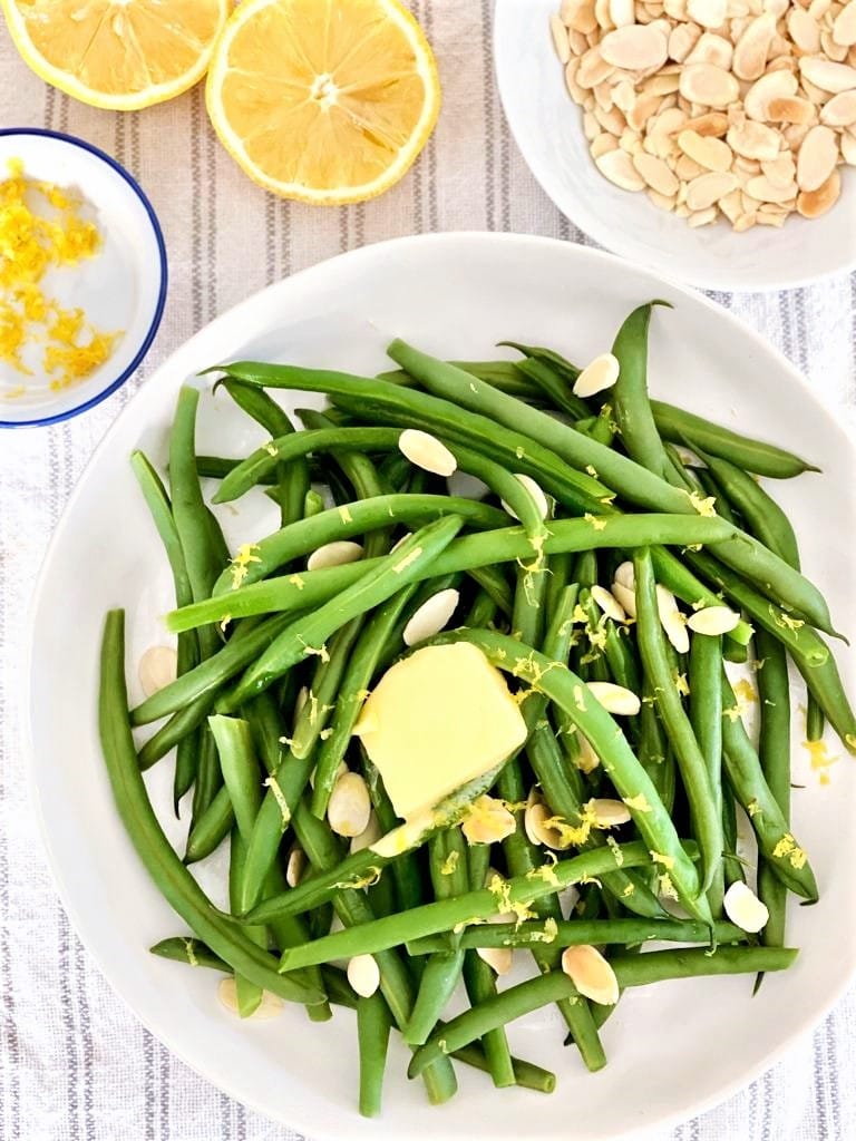 White bowl with cooked green beans, slivered almonds, lemon zest and a knob of butter with separate bowls with recipe ingredients set alongside