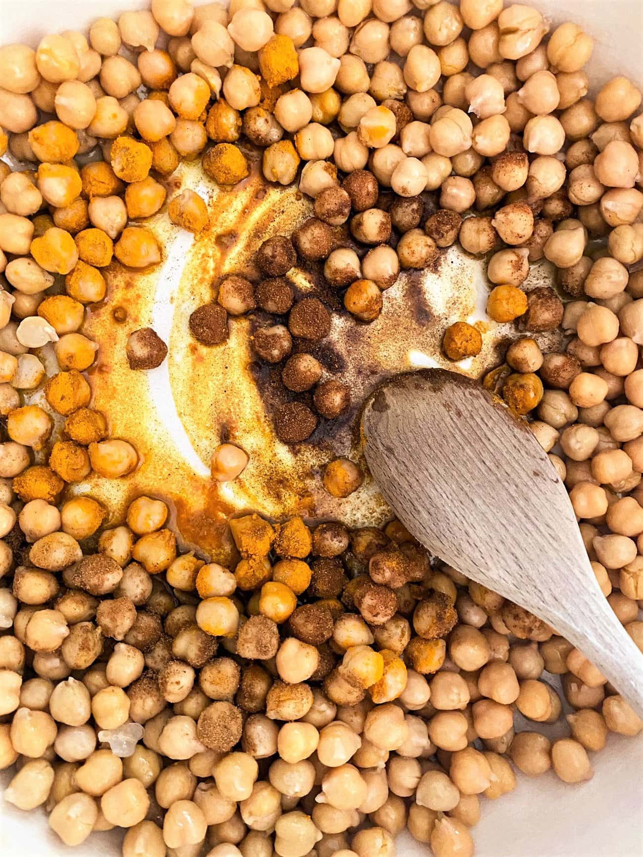 Cast iron skillet with drained chickpeas covered in blend of oil and spices with a wooden spoon