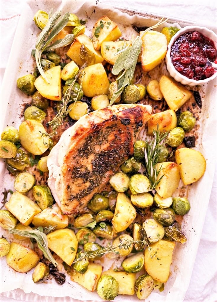 A sheet tray filled with roasted potatoes and brussels sprouts, sprigs of rosemary, thyme and sage with a whole bone-in, skin-on honey glazed turkey breast set on top of the vegetables and a top of cranberry sauce