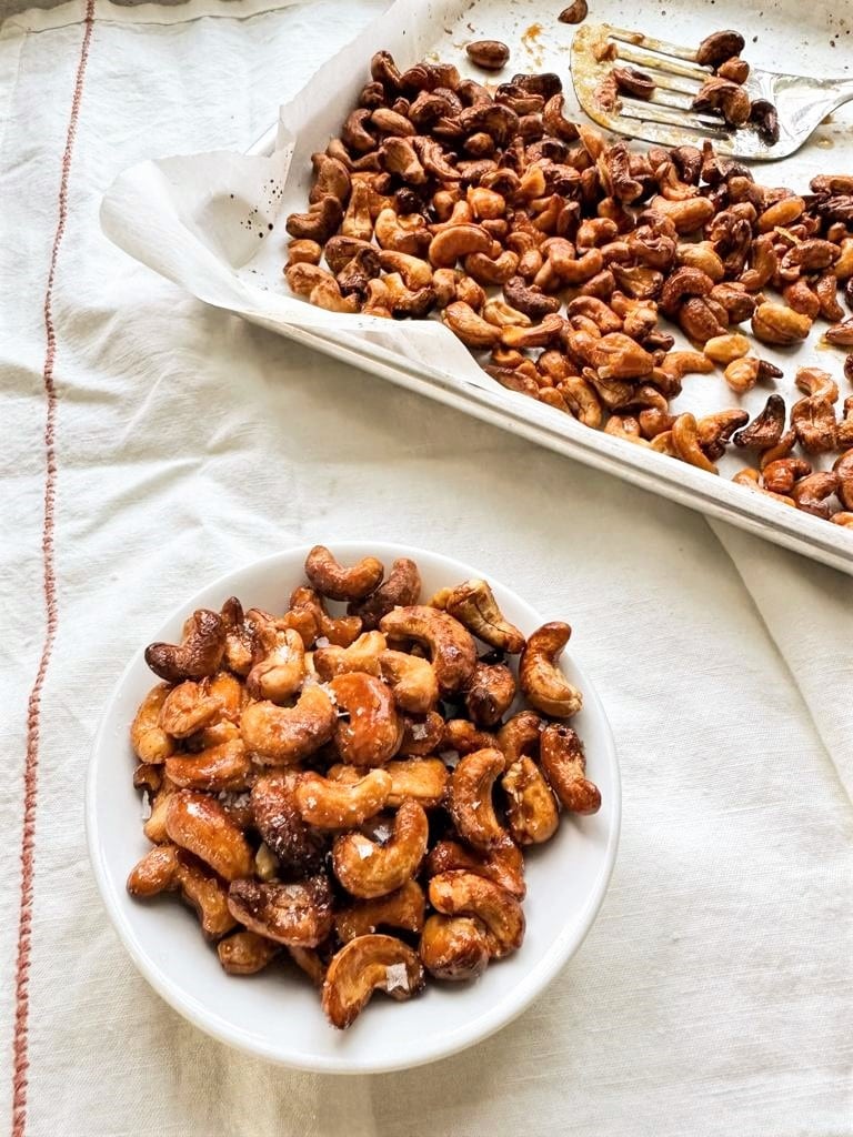 A white bowl filled with honey roasted cashews and a paper lined baking sheet filled with honey roasted cashews, and a metal spatula set alongside.