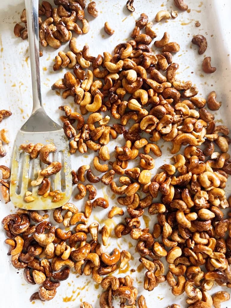 Paper lined baking sheet filled with honey roasted cashews, with a metal spatula set alongside.