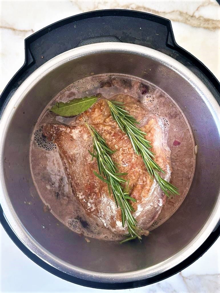 Instant pot bowl filled with broth and boneless leg of lamb, topped with bay leaves and 2 sprigs of fresh rosemary