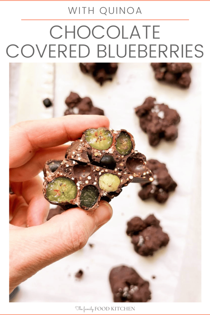 A pinnable image with recipe title and photo of hand holding a chocolate covered blueberry cluster cut in half and a paper lined baking sheet covered with clusters of chocolate covered blueberries in the background.