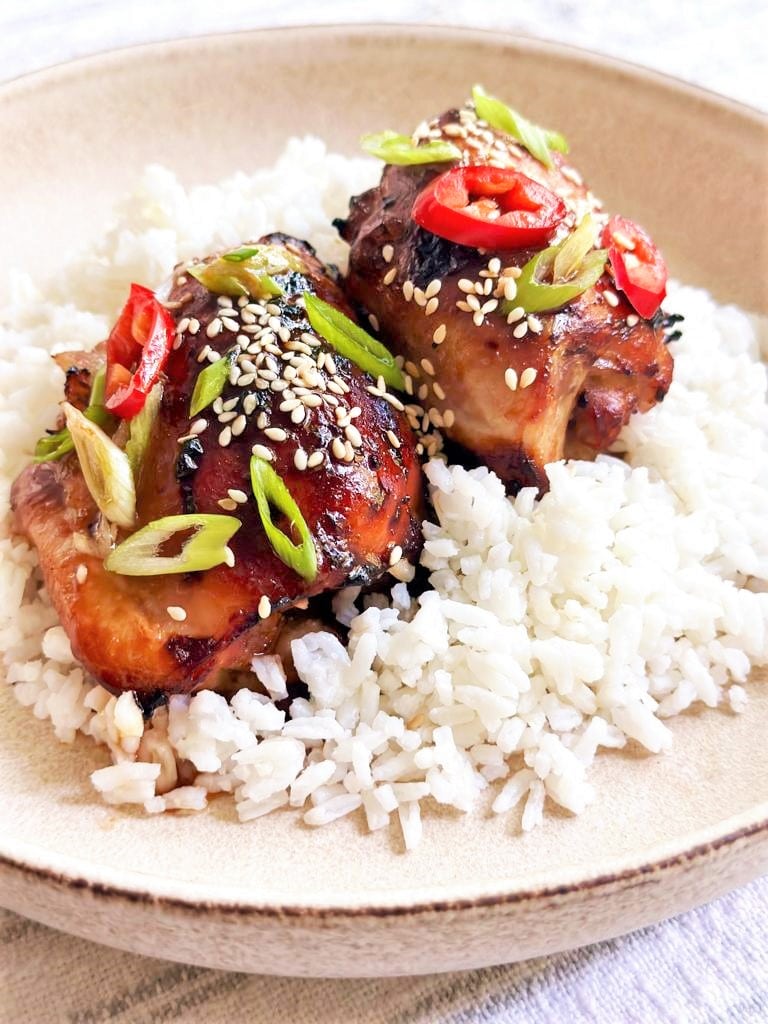 Bowl filled with steamed white rice topped with 2 miso glazed chicken thighs garnished with sesame seeds, sliced green onion and red chili.