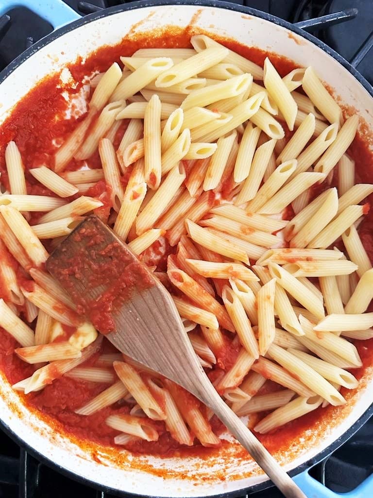 Dutch oven filled with tomato sauce and topped with freshly cooked pasta and a wooden spatula.