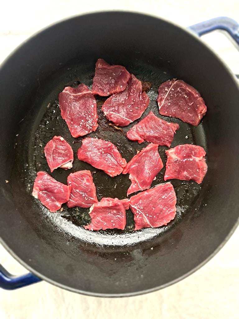 Dutch oven with thin slices of beef being seared in a single layer.