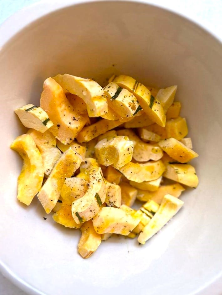 A bowl filled with slices of delicata squash tossed in olive oil and seasoned with salt and black pepper.