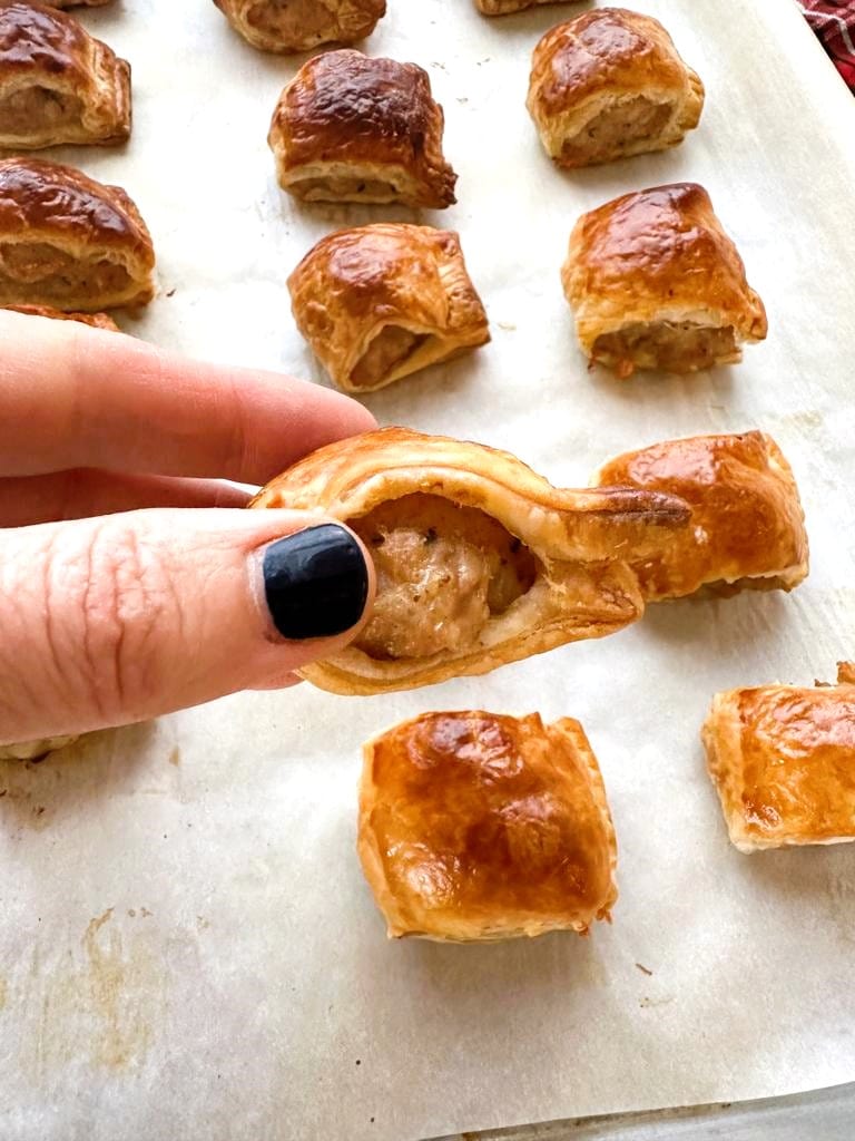 Cooked chicken sausage rolls set out on a sheet of baking parchment with a hand holding a close up image of a sausage roll.