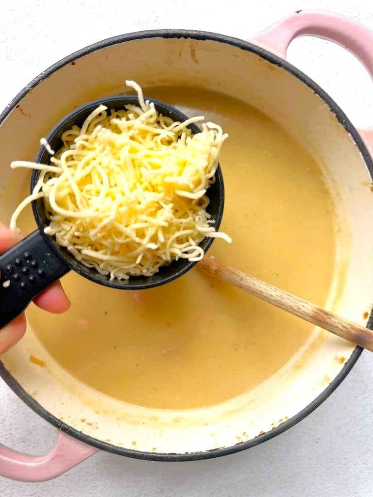 A cup of shredded cheddar cheese being added to a Dutch oven filled with creamy leek and potato soup.