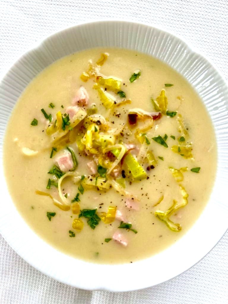 A white bowl filled with creamy leek, ham & potato soup garnished with sauteed leeks and cubes of cooked ham.