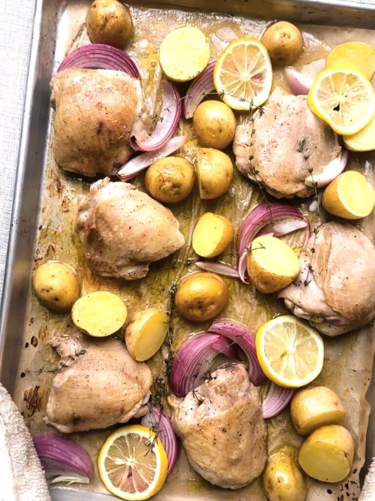 Part roasted bone-in, skin-on chicken thighs, sliced red onions and halved baby potatoes spread over a parchment lined sheet pan.
