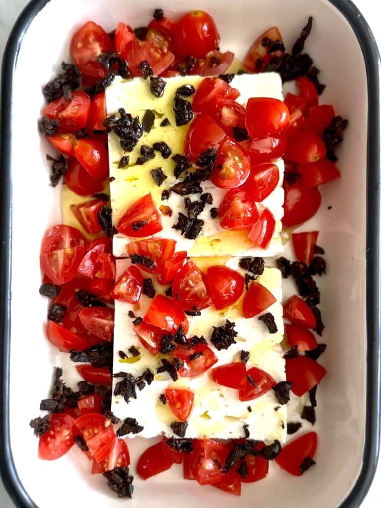 A shallow dish with 2 blocks of feta cheese, topped with fresh cherry tomatoes and chopped sundried tomatoes, with a drizzle of olive oil.