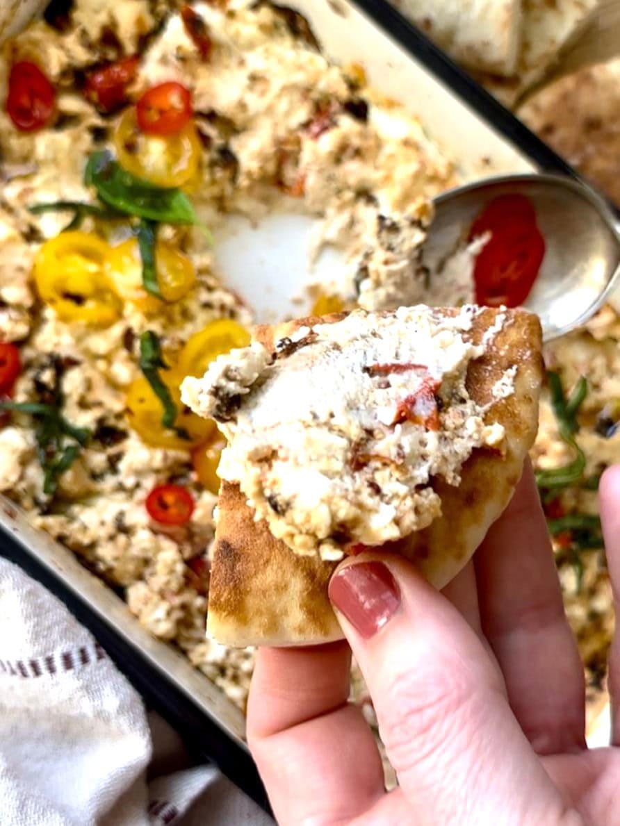 A shallow dish of hot baked feta cheese dip, topped with fresh tomatoes and shredded basil leaves, with a hand holding a wedge of pita bread topped with feta cheese dip.