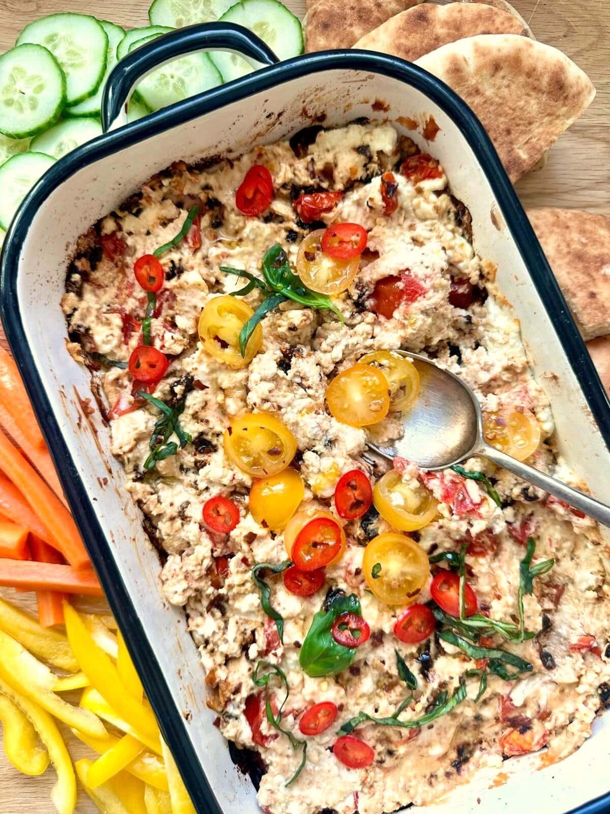 A shallow dish of hot baked feta cheese dip, topped with fresh tomatoes and shredded basil leaves, served with pita wedges and vegetable sticks set alongside.