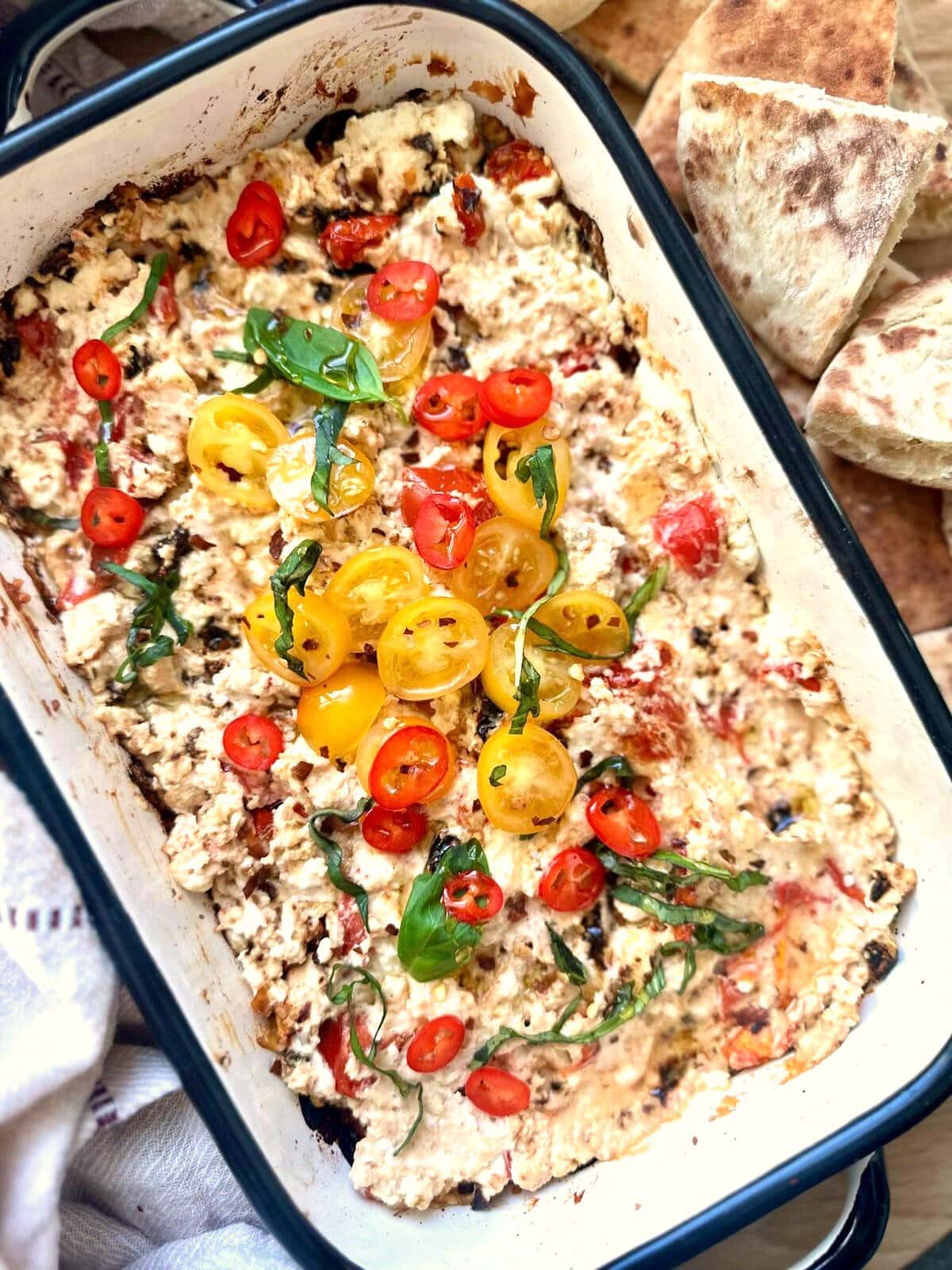 A shallow dish of hot baked feta cheese dip, topped with fresh tomatoes and shredded basil leaves.